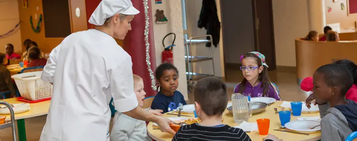une cantine scolaire avec des enfants servis par une employée