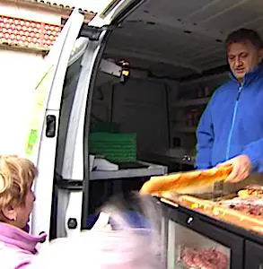 Un commerçant ambulant, dans son camion, qui vend une baguette de pain à une cliente. La renaissance des commerces ambulants s'adapte aux besoins du monde rural.